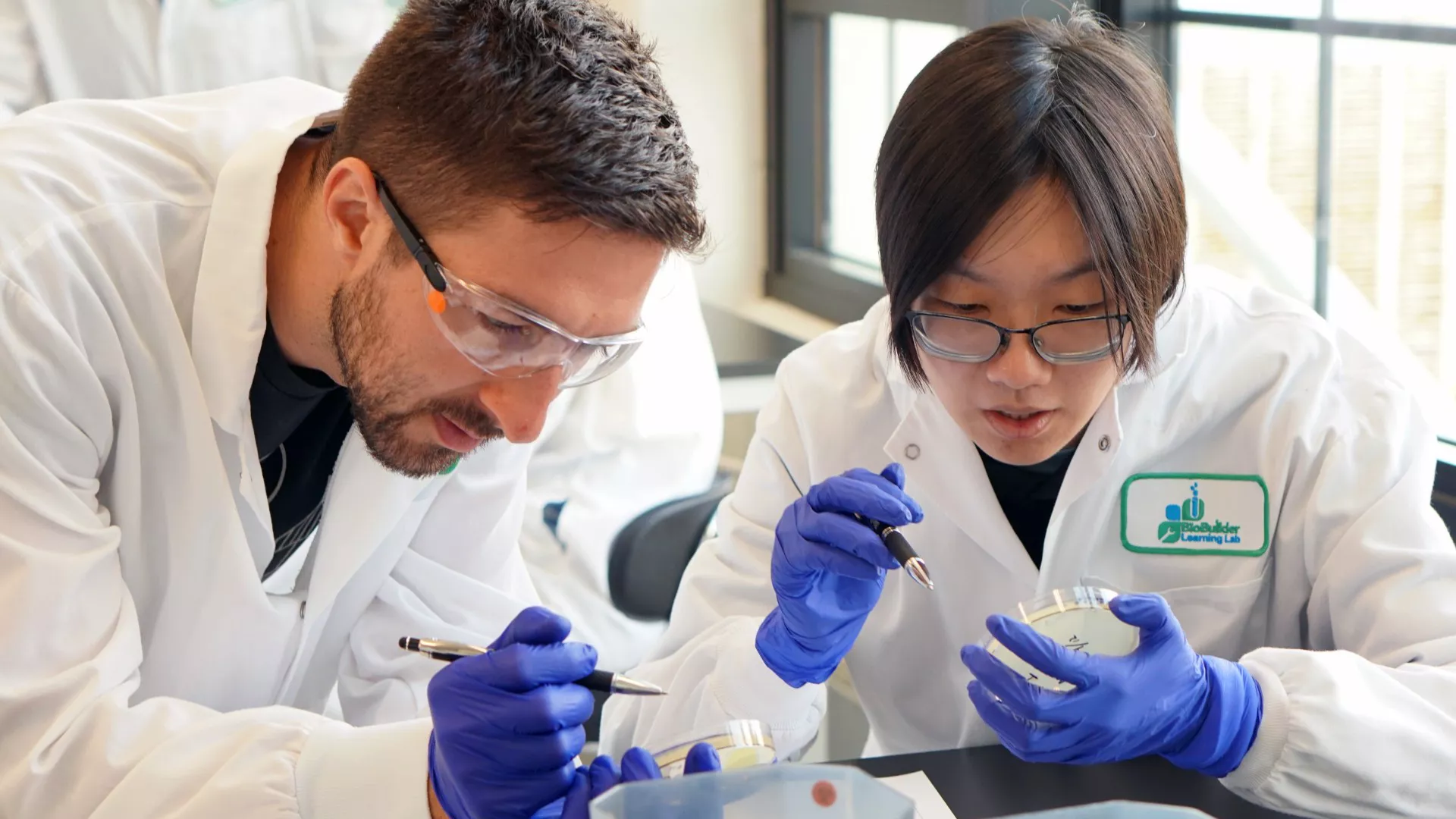High school student and scientist examine petri dishes while wearing labcoats, gloves, and googles