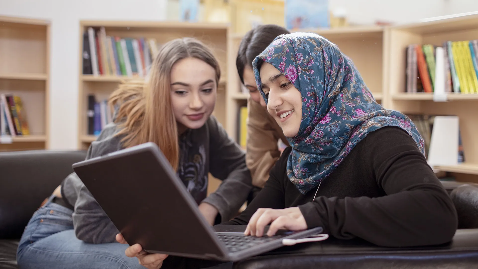 Three high school students, one wearing a headscarf, work on a laptop together