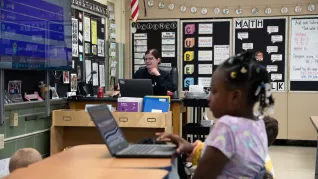 Ms. Tingle sits at her classroom desk while her student works on her laptop in the foreground