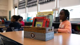 closeup photo of a rectangular wood and metal caddie on a students' desktop. Caddie has book, folders, and pencil case showing.
