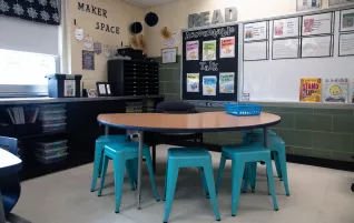 corner of an elementary classroom with information hanging on the whiteboard and a table with 5 teal stools