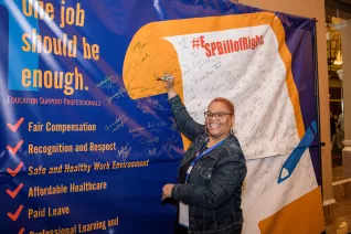 a photo of an education support professional holding a pen up to sign a large yellow-outlined scroll of the ESP Bill of Rights.
