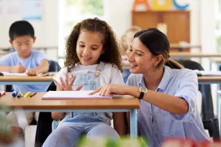 Female students sitting at a desk, with a teacher kneeling at her side.