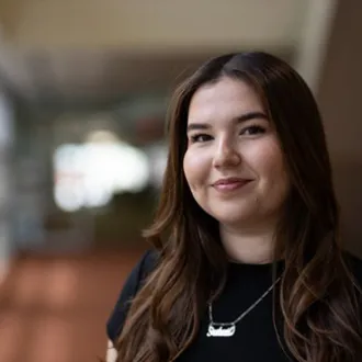 A woman standing in a blurred hallway and smiling softly.